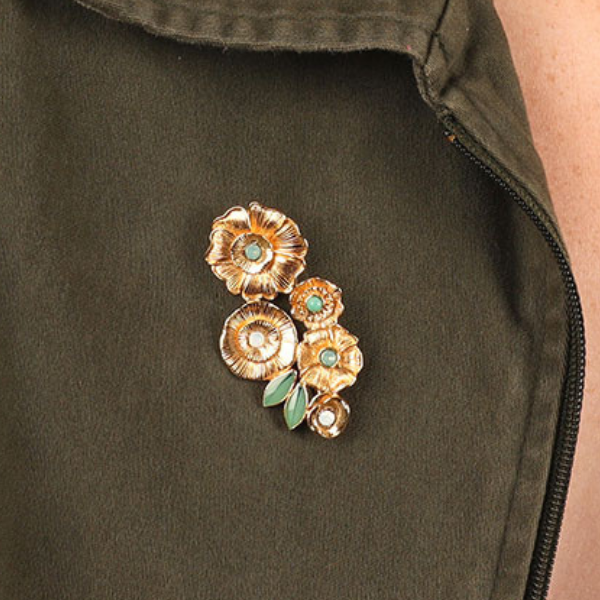 Image of model wearing golden brooch with four different shaped flowers with blue green rhinestone in centre of each on gold metal finish.