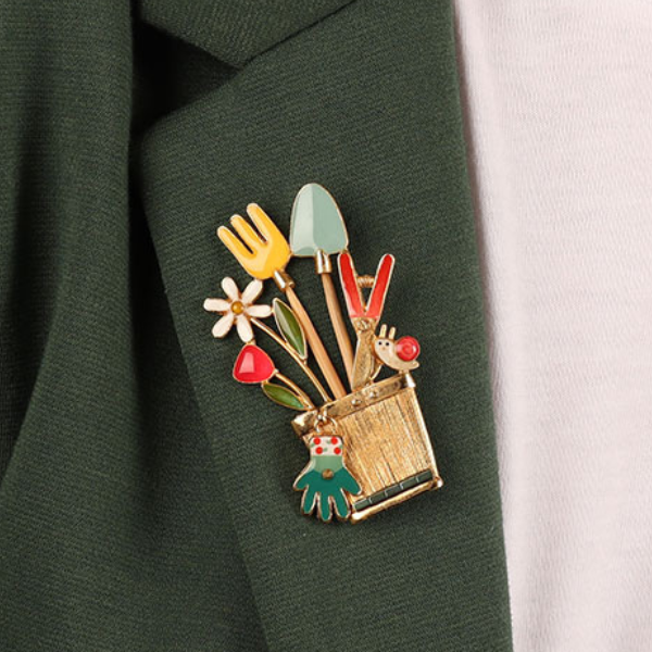 Image of model wearing quirky brooch featuring garden tools and flowers all hand painted using precious beads and stones on gold plated finish.