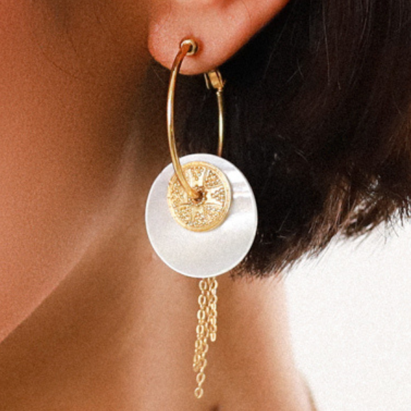 Image of model wearing creole earrings with mother of pearl disc and gold metal tassels dangle.