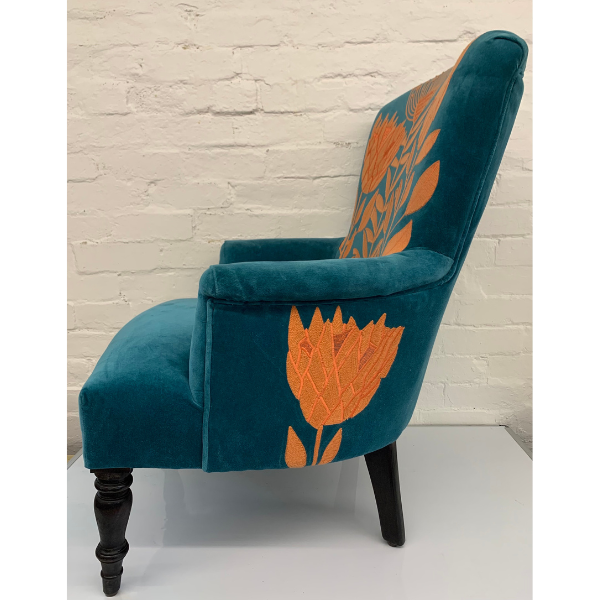 Image of the side view of a velvet turquoise arm chair with orange waratah embroidery.